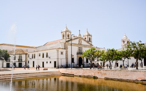 Blick auf die weiße Kirche mit zwei Glockentürmen neben einem Platz und einem flachen Becken in Faro