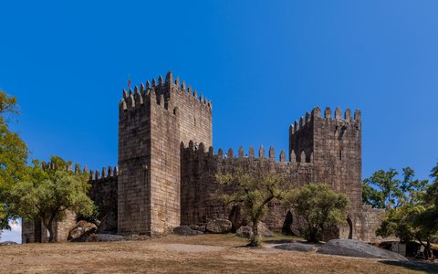Der steinerne Bergfried und die Zinnen der Burg von Guimarães zwischen Bäumen vor klarem blauem Himmel