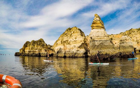 Goldene Sandsteinfelsen und Klippen bei Lagos, die sich im ruhigen grünblauen Wasser spiegeln, mit Stand-up-Paddlern und bewölktem blauem Himmel