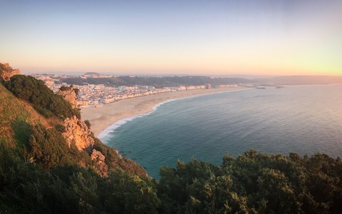 Der lange Strand und die Stadt Nazaré, die sich im warmen Abendlicht an der Bucht entlangziehen, gesehen von der Klippe