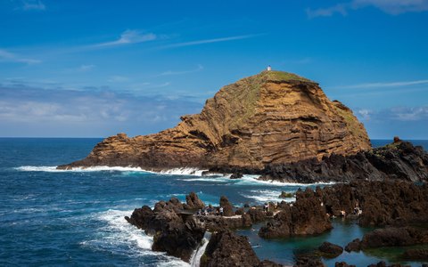 Eine kleine Felseninsel mit einem kleinen Leuchtturm vor der Lavaküste von Porto Moniz, mit Wellen und blauem Himmel