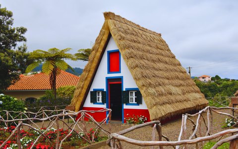 Ein dreieckiges, weiß, rot und blau bemaltes Santana-Haus mit Strohdach, davor Baumfarne und Blumen