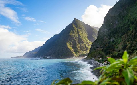 Steile grüne Berge, die zu einer felsigen Küste und blauem Meer bei São Vicente an der Nordküste Madeiras abfallen, unter blauem Himmel mit weißen Wolken