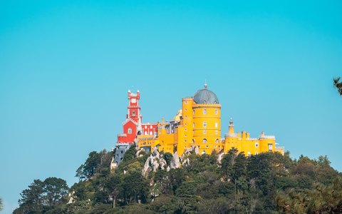 Die roten und gelben Türme des Palácio da Pena auf der Kuppe eines bewaldeten Hügels vor klarem blauem Himmel