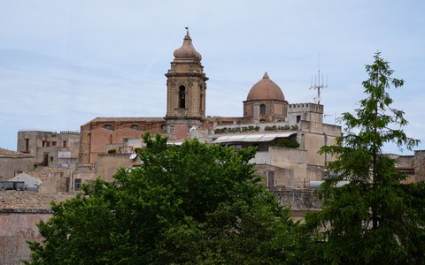 Alter steinerner Kirchturm und Dächer von Erice hinter grünen Bäumen unter blauem Himmel mit weißen Wolken