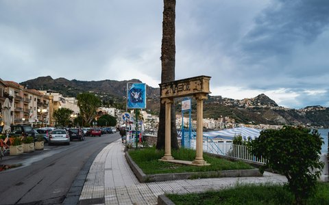 Uferstraße in Giardini Naxos mit Palmen und Steinsäule unter bewölktem Himmel, dahinter die Hügel von Taormina