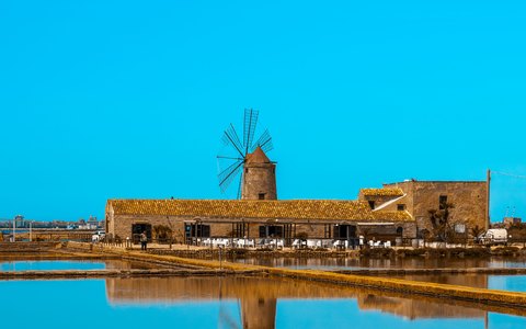 Steinerne Windmühle und Salinen spiegeln sich unter strahlend blauem Himmel im stillen Wasser