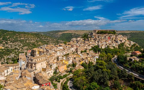 Luftaufnahme der alten Hügelstadt Ragusa Ibla mit Kuppeln zwischen grünen Hügeln unter blauem Himmel
