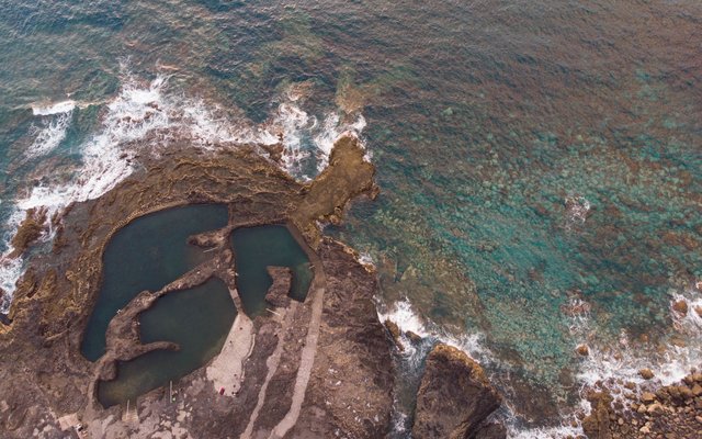 Luftaufnahme dunkler Felsbecken auf einer felsigen Landzunge bei Agaete, mit brechenden Wellen entlang der Küste und blaugrünem Meer ringsum