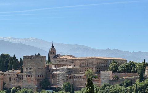 Blick auf die Alhambra in Granada, mit Mauern und Türmen über grünen Bäumen und der Sierra Nevada dahinter