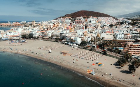 Luftbild des Sandstrands und des weißen Orts Los Cristianos unter einem braunen Hügel am blauen Meer