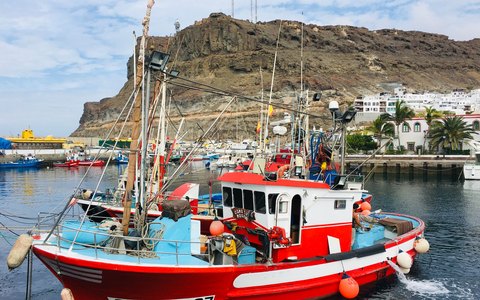 Ein rot-weißes Fischerboot liegt im Hafen von Mogán unterhalb eines felsigen Hügels unter blauem Himmel vertäut