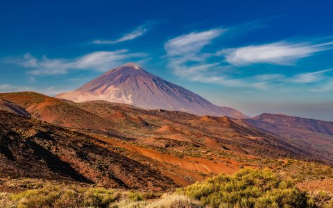 Teide-Gipfel über rotem und braunem Vulkanboden unter tiefblauem Himmel mit weißen Wolken