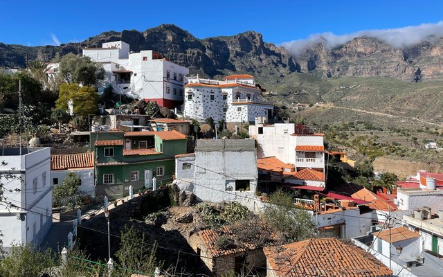 Weiße und bunte Häuser mit Ziegeldächern an einem Hang in Tejeda, unterhalb zerklüfteter Berge und unter blauem Himmel