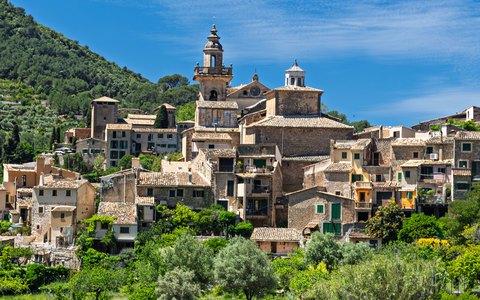 Das Dorf Valldemossa mit seinen Steinhäusern und seinem Kirchturm zwischen grünen Bäumen unter blauem Himmel