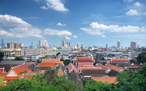 Traditionelle thailändische Tempeldächer vor der modernen Skyline Bangkoks unter blauem Himmel mit weißen Wolken