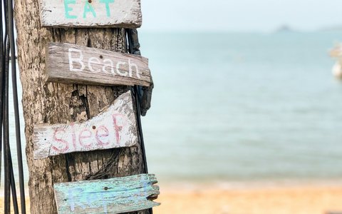 Handbemalte Holzschilder mit den Wörtern Eat, Beach und Sleep an einem Baumstamm, dahinter Sand und ruhiges Meer am Bophut Beach