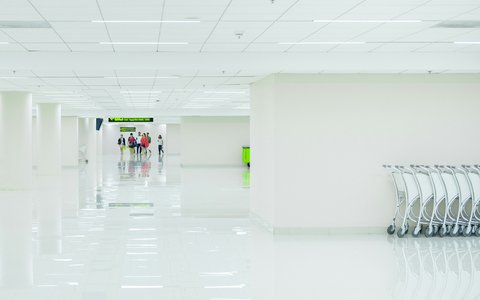 Langer, heller weißer Terminalgang am Flughafen Don Mueang, rechts aufgereihte Gepäckwagen und einige Reisende, die auf ein Schild zugehen