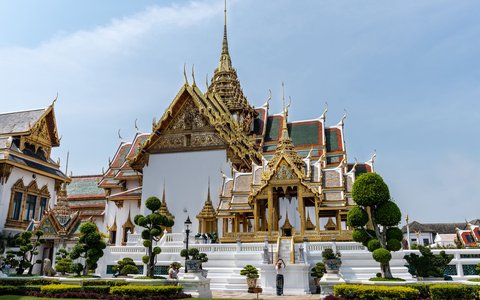 Blick auf die vergoldeten Türme und weißen Hallen des Großen Palastes in Bangkok hinter gestutzten Bäumen und Rasen