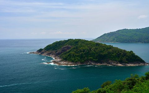 Unter hellblauem Himmel eine grüne, bewaldete Insel im glitzernden blauen Meer vor der Küste bei Kamala