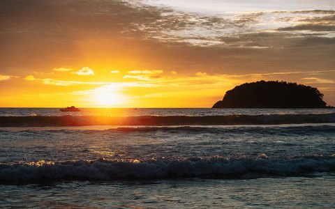 Die Sonne geht hinter einer kleinen Insel vor dem Kata Beach unter, Wellen rollen unter einem goldenen Himmel heran
