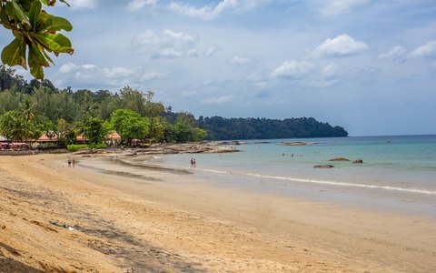 Ein ruhiger Sandstrand, der sich in Khao Lak um eine Bucht schwingt, mit Palmen, grünen Hügeln und blauem Himmel mit Wolken