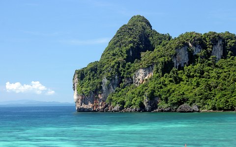 Unter blauem Himmel erhebt sich eine mit grünem Dschungel bewachsene Kalksteinklippe aus türkisfarbenem Wasser bei Ao Nang