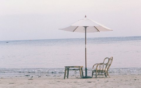 Ein weißer Sonnenschirm über einem Holzstuhl und einem kleinen Tisch im Sand am Lamai Beach, dahinter ruhiges Meer und ein blasser Himmel