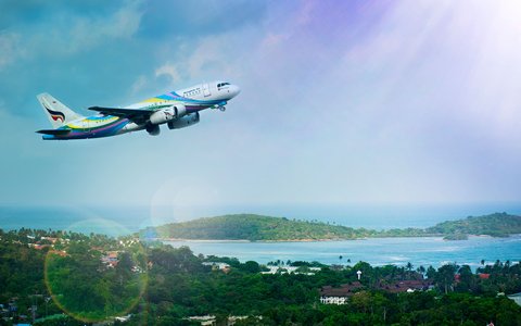 Buntes Verkehrsflugzeug steigt über grünen Hügeln und einer Bucht auf Koh Samui auf