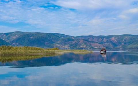 Ruhiger See mit Spiegelungen von Schilf und bewaldeten Hügeln bei Dalyan unter blauem Himmel mit weißen Wolken