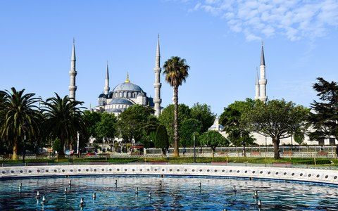 Kuppeln und Minarette der Blauen Moschee hinter Gärten, Palmen und einem Brunnen unter blauem Himmel