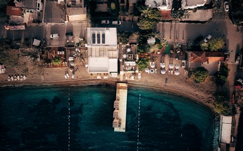 Drohnenaufnahme von oben auf eine kleine Bucht in Yalıkavak mit Steg, Sandstrand mit Sonnenschirmen und Gebäuden am Ufer