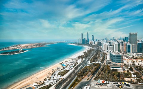 Luftaufnahme der Corniche von Abu Dhabi mit Strand, türkisfarbenem Wasser und Hochhäusern unter blauem Himmel mit Wolkenschleiern