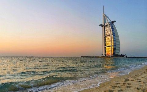 Blick vom Jumeirah Beach auf das segelförmige Burj Al Arab am Meer, mit kleinen Wellen und einem Himmel im Licht des Sonnenuntergangs